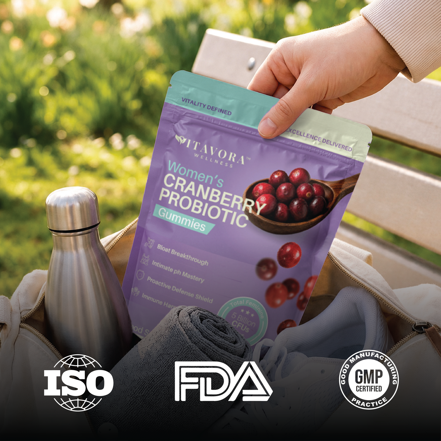 A lifestyle product image showing a hand lifting a purple Vitavora Women's Cranberry Probiotic Gummies pouch out of a canvas gym bag next to a stainless steel water bottle, completed with trust badges for ISO, FDA registered facility, and GMP Certified Practice.
